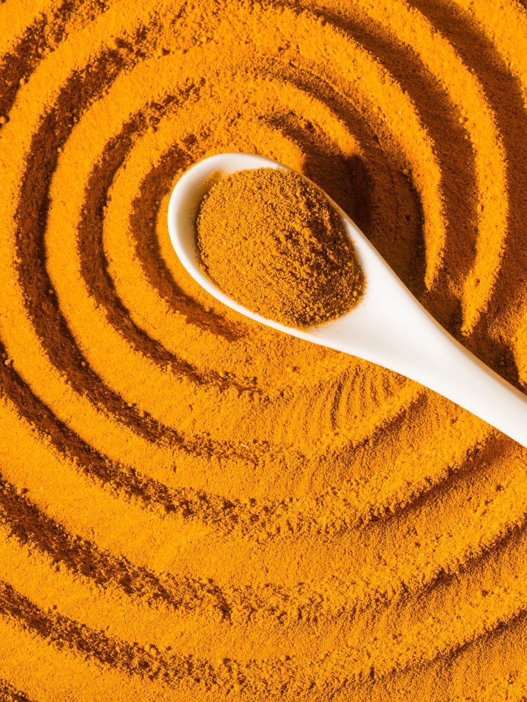 A white spoon of golden turmeric powder on a swirled bed of turmeric, the spice behind the salve's turmeric butter