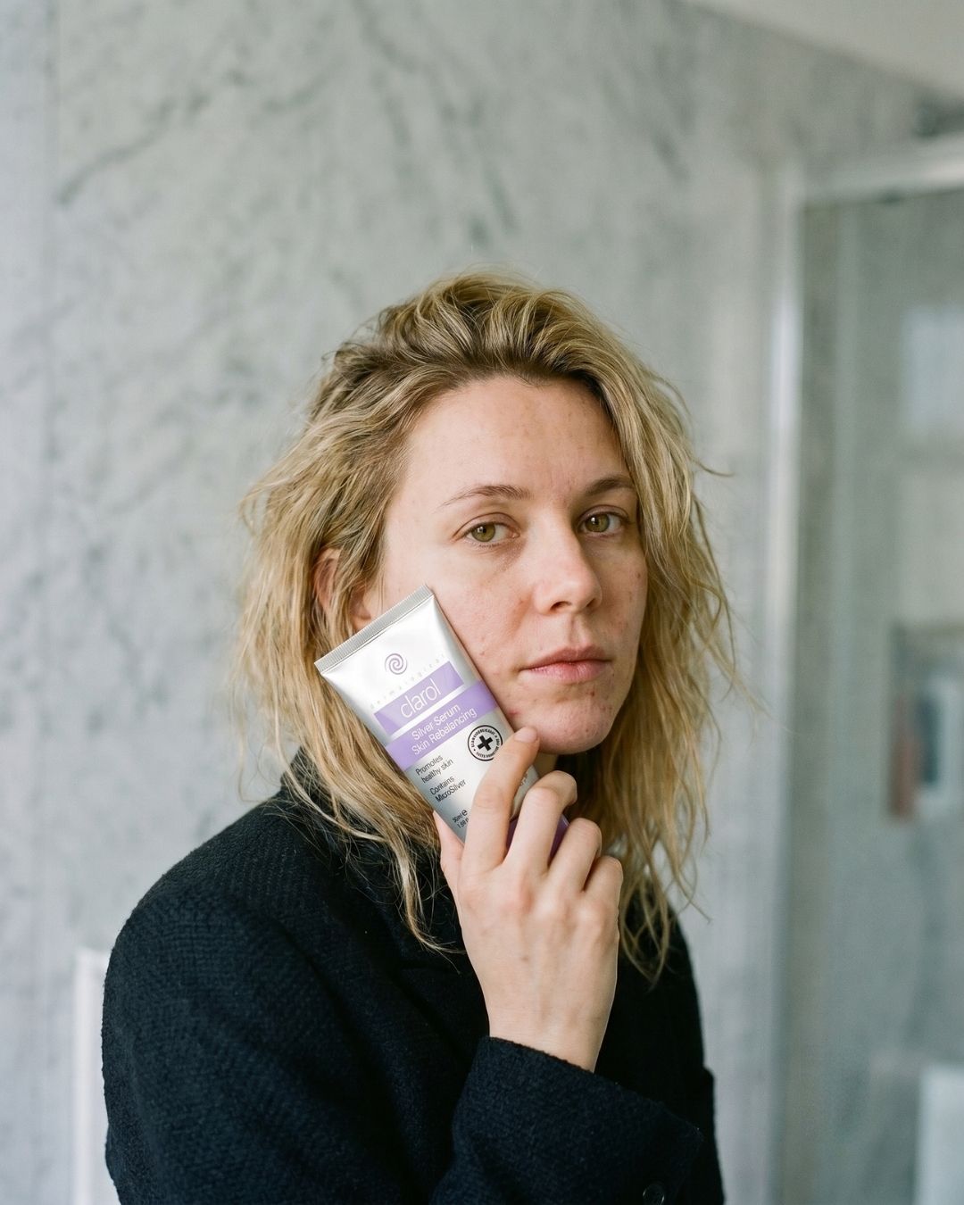 Woman with spot-prone skin holding a tube of Clarol Silver Serum Skin Rebalancing with MicroSilver to her cheek as part of her daily acne skincare routine