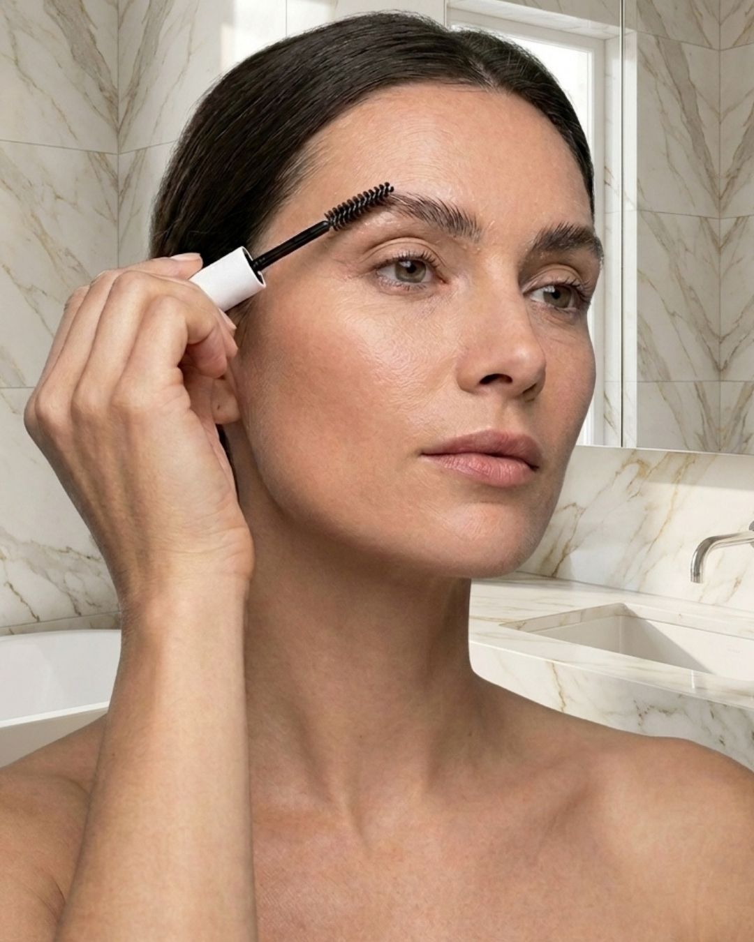 A woman applying Hydrosil Hydrobrow Serum to her eyebrow with the brush applicator, in a bathroom