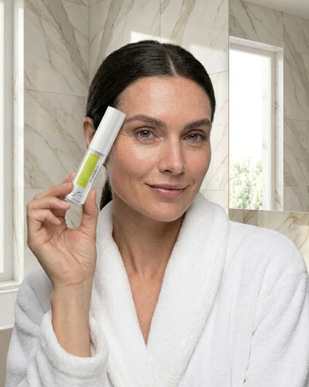 A woman in a white robe holding up a bottle of Hydrosil Hydrobrow Serum beside her brows, in a bathroom