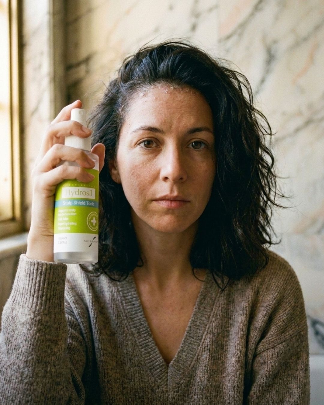 A woman with dark, wavy hair looks at the camera, holding a bottle of Hydrosil Scalp Shield Tonic up beside her face with the green label facing forward.