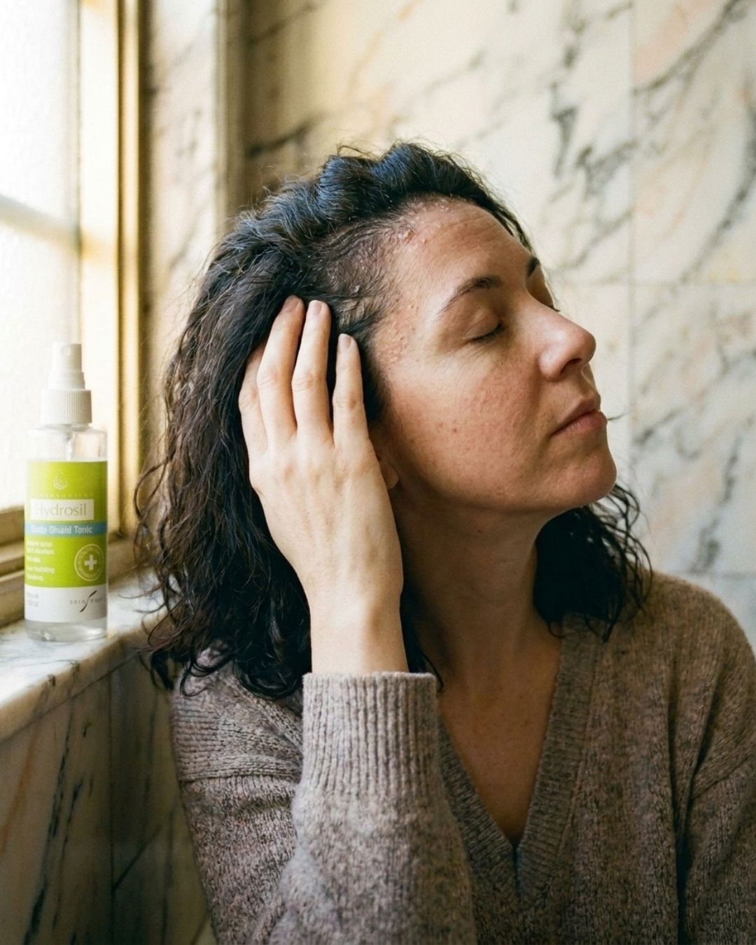 A woman with dark, wavy damp hair sits by a window, eyes closed, massaging her scalp with her fingertips, a bottle of Hydrosil Scalp Shield Tonic on the sill.
