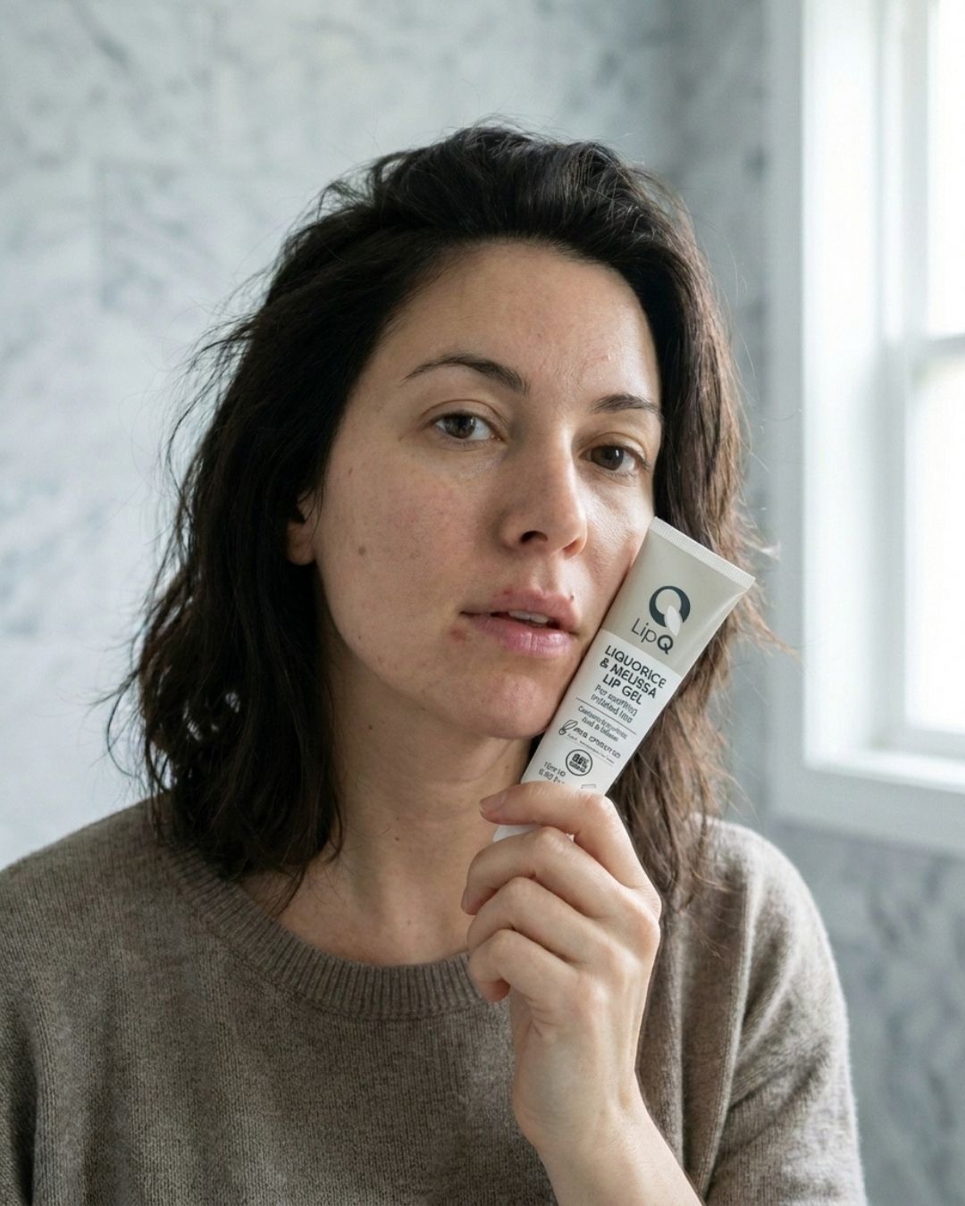 A woman holding a tube of LipQ Liquorice & Melissa Lip Gel beside her face, a lip gel for cold sore-prone lips